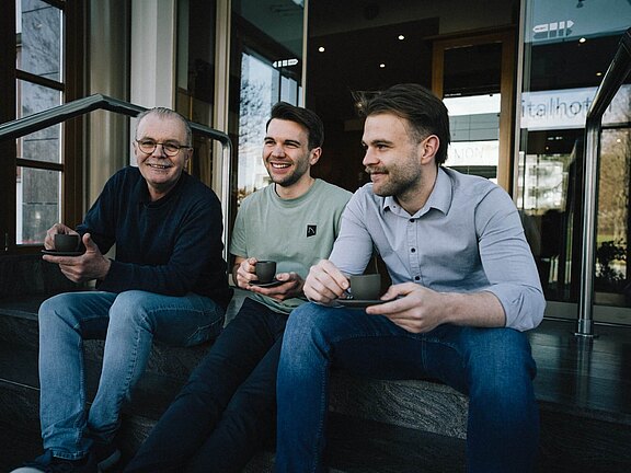 Drei Generationen der Gastgeberfamilie Simon sitzen entspannt vor dem Hotel und genießen gemeinsam eine Tasse Kaffee.